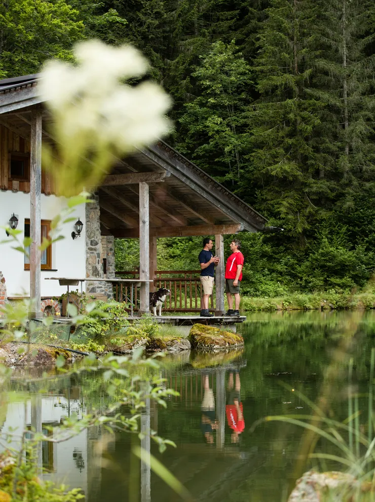 Two men talking on a cabin porch by a forest lake with a dog nearby