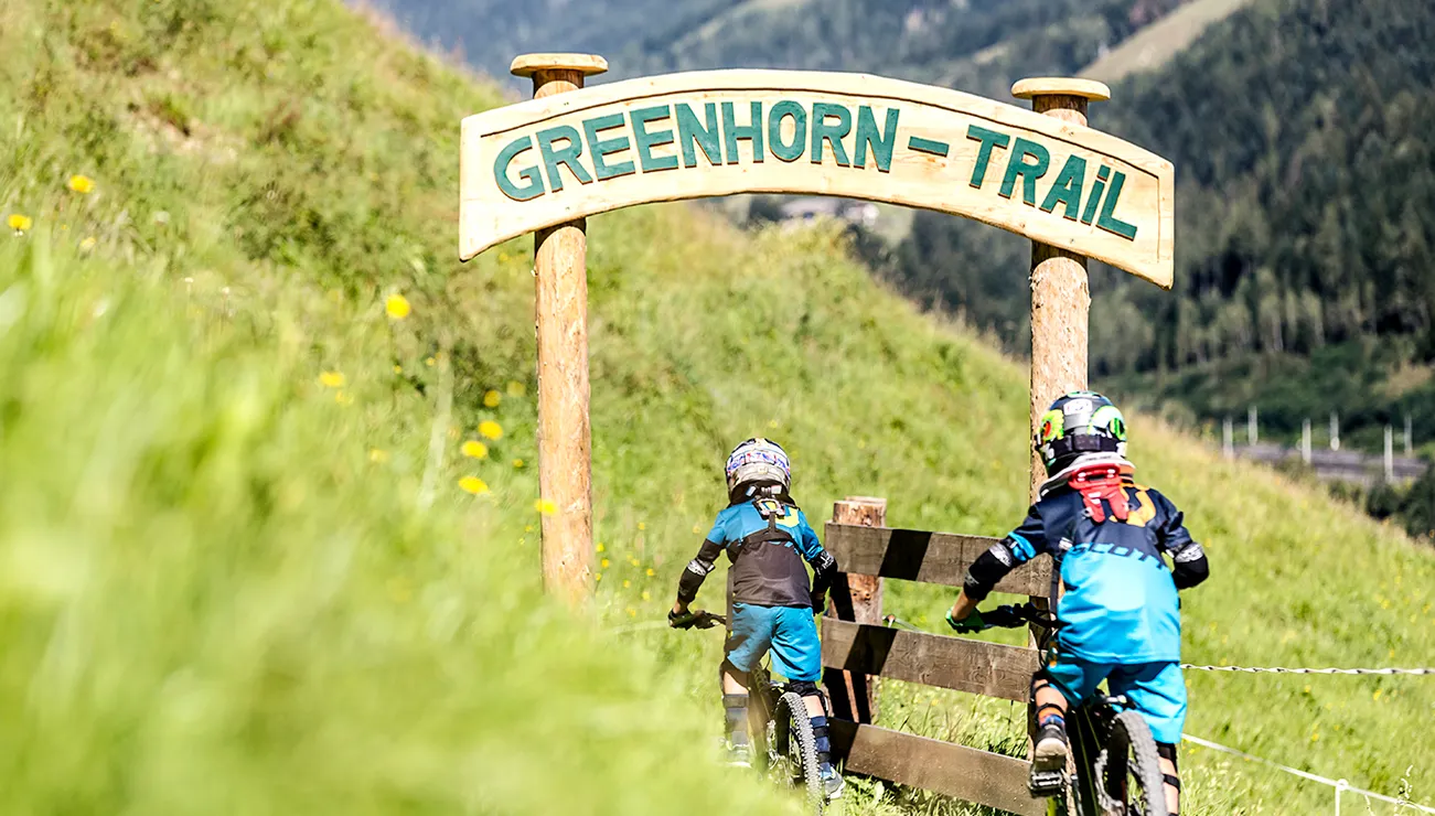 Two kids on mountain bikes entering Greenhorn Trail in a grassy alpine landscape