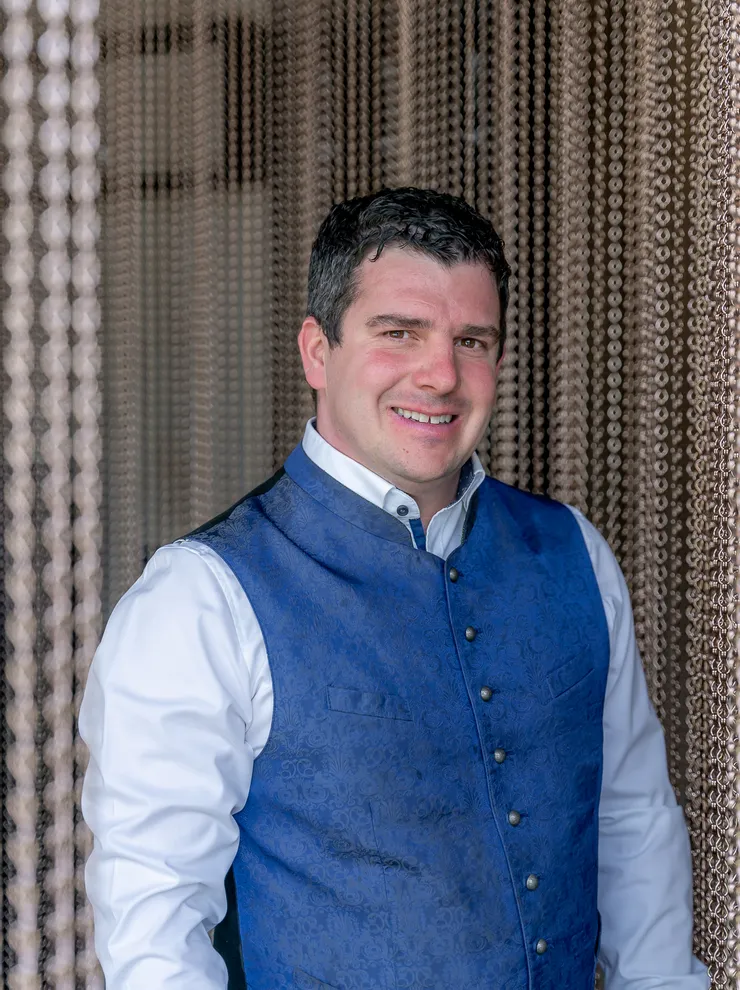 Man wearing traditional blue vest standing in front of beaded curtain