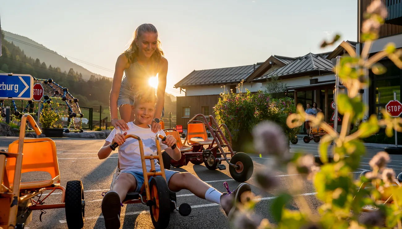 Boy riding a go-kart in the sunshine with girl pushing behind in a fun outdoor setting