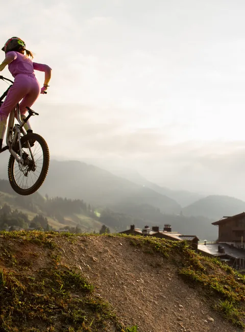Mountain biker in pink gear doing a jump on dirt trail at sunset near alpine village