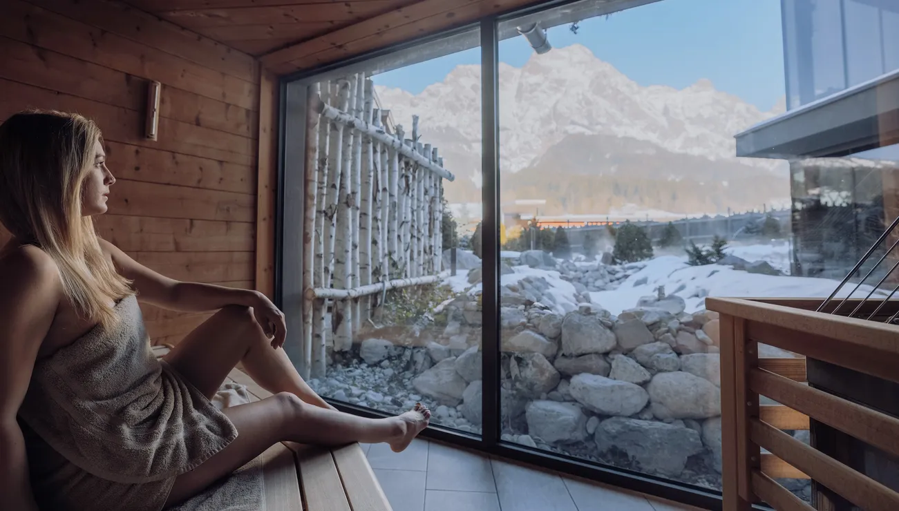 Woman relaxing in sauna with scenic snowy mountain view through large glass window
