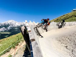 Mountain bikers riding on a steep wooden berm in an alpine bike park with mountain views