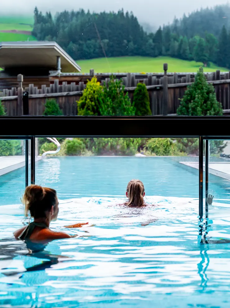 Two women swimming in an infinity pool overlooking green hills and forest