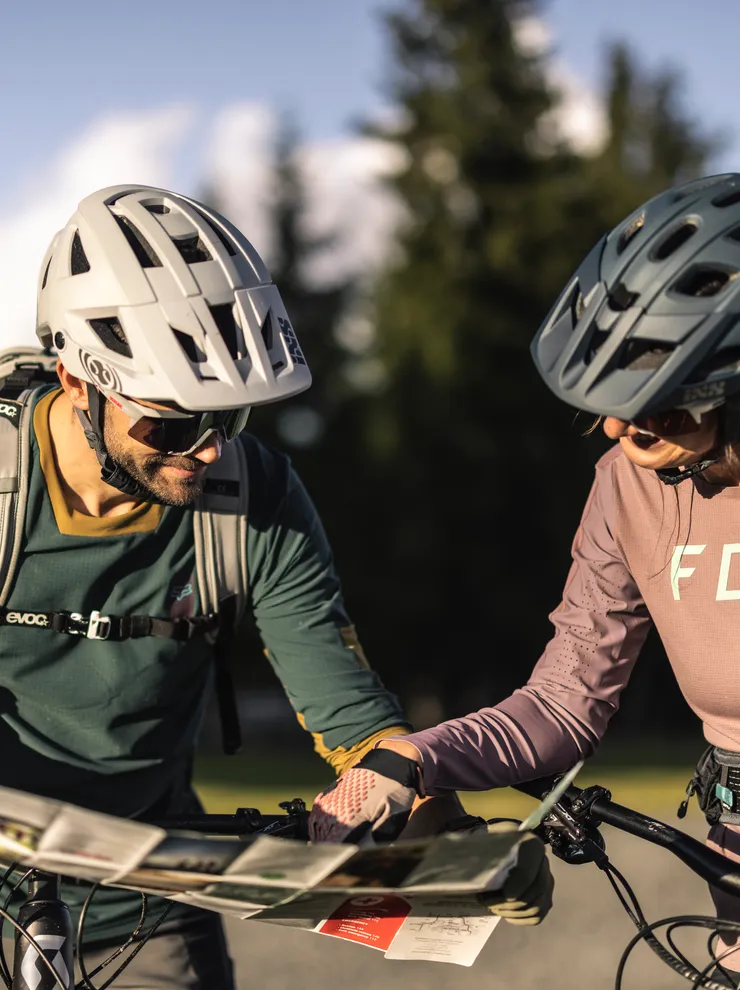 Two mountain bikers in helmets reading a map on a sunny trail with alpine scenery