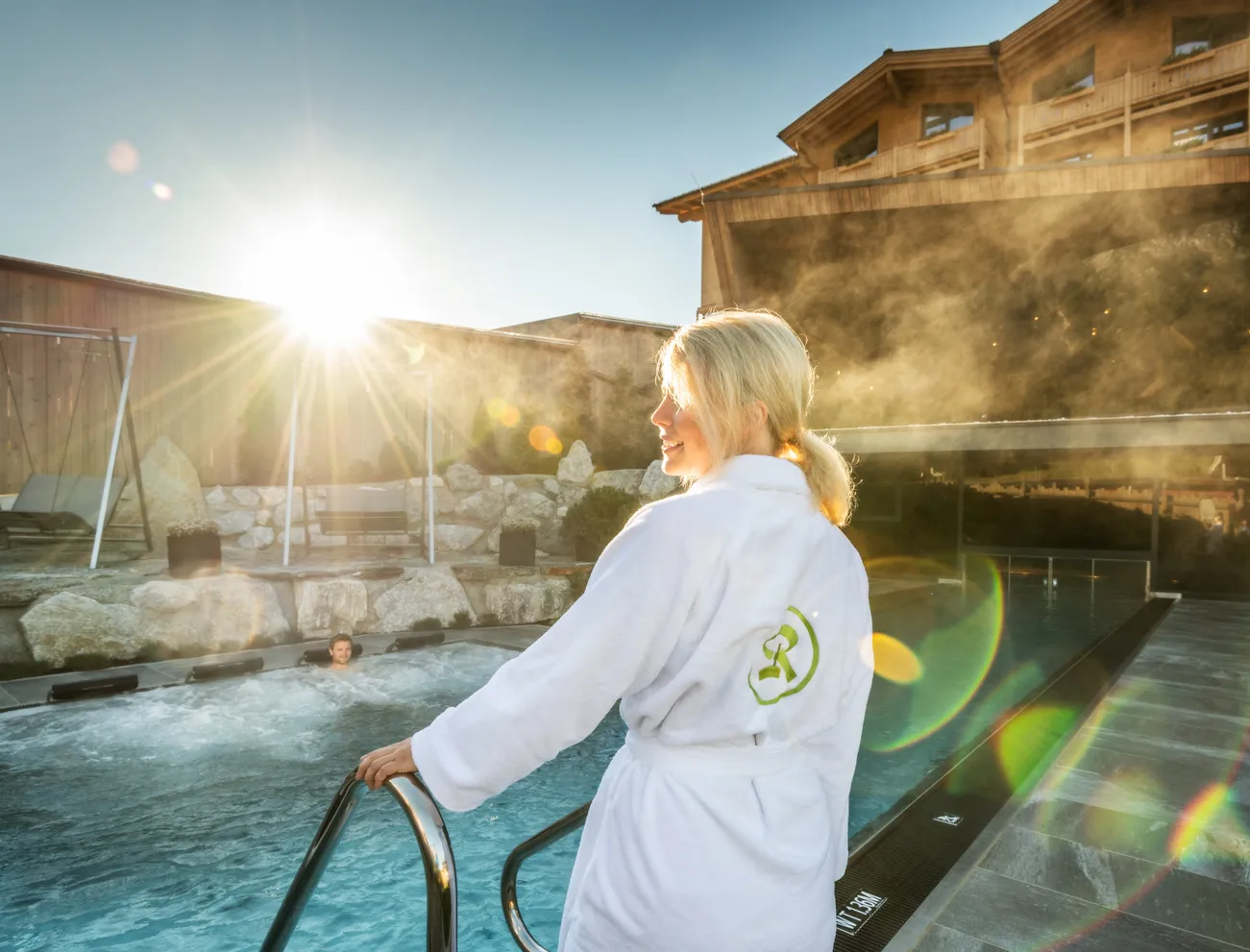 Frau im weißen Bademantel steigt in Außenpool eines Luxus-Wellnessresorts