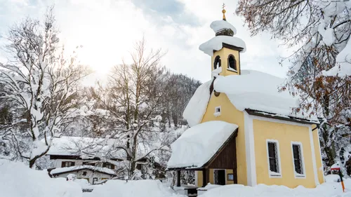 Verschneite gelbe Kapelle in winterlicher Berglandschaft mit Bäumen und Dorfhäusern