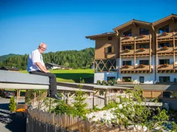 Mann sitzt am Hoteldachrand vor alpinem Holzgebäude und Waldkulisse