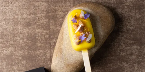 Gourmet-Eis am Stiel mit gelbem Überzug, essbaren Blumen und Nüssen auf Stein neben Safran-Box