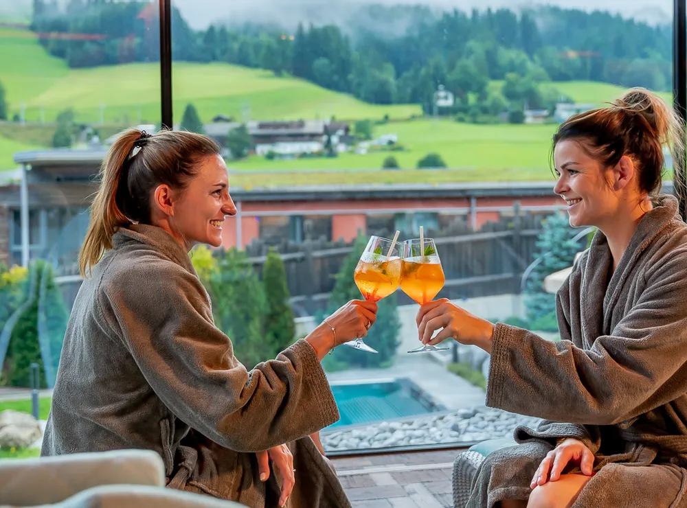 Zwei Frauen im Bademantel stoßen mit Getränken im Wellnessresort mit Bergblick an