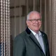 Smiling older man in a green blazer with glasses standing in front of a curtain of metal chains