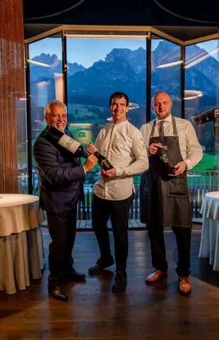 Three men in fine dining restaurant holding wine bottles with mountain view in background