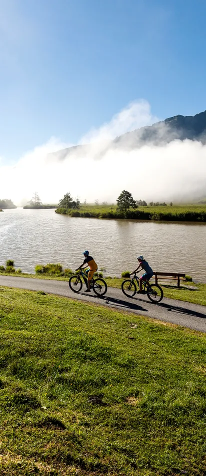 Cyclists riding beside mountain lake on a sunny morning with misty hills