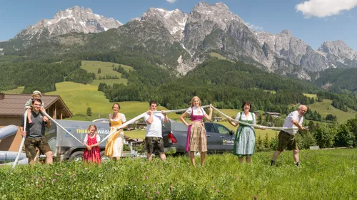 Menschen in traditioneller österreichischer Kleidung mit Schlauch auf Wiese vor Alpenpanorama