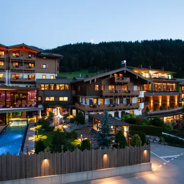 Beleuchtetes Alpenhotel mit Außenpool und Balkonen bei Sonnenuntergang
