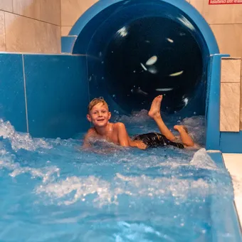 Junge rutscht lachend aus einer blauen Wasserrutsche in einem Hallenbad