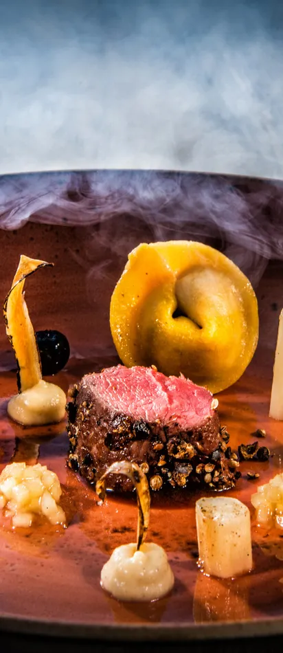 Gourmet-Rindfleischgericht mit Gemüse, Ravioli, Soße und Rauch auf rotbraunem Keramikteller