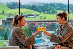 Zwei Frauen im Bademantel stoßen mit Getränken im Wellnessresort mit Bergblick an