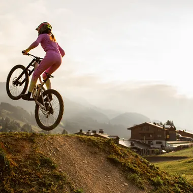 Woman in pink outfit mountain biking over dirt ramp at sunset