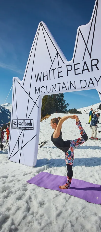 Frau macht Yoga im Schnee bei den White Pearl Mountain Days in Saalbach mit Skifahrern im Hintergrund