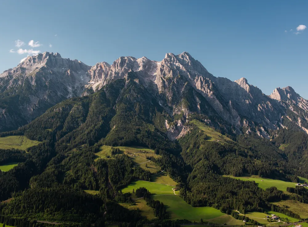 Alpine mountain range with forested slopes and rocky peaks under clear blue sky