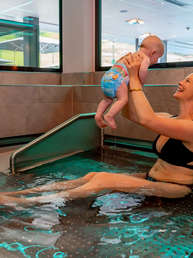 Smiling mother in bikini lifts baby in swim diaper in indoor pool