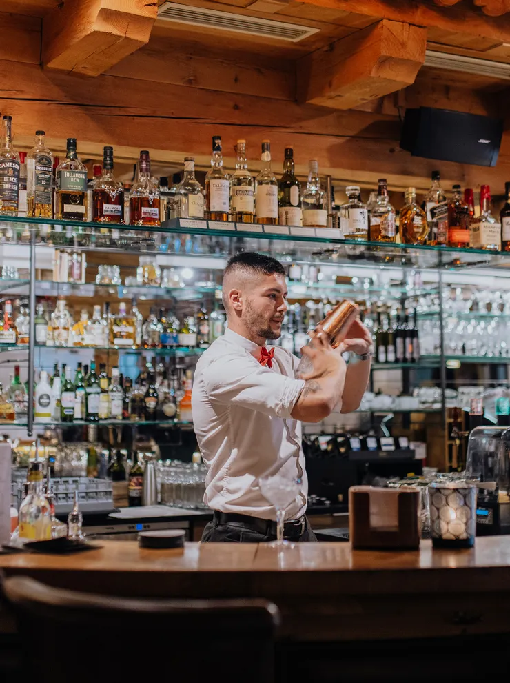 Barkeeper schüttelt Cocktail an Holzbar vor Regal mit Spirituosenflaschen