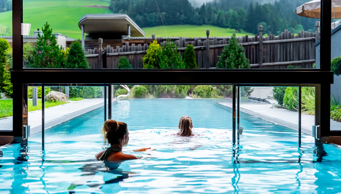 Two women swimming in an infinity pool overlooking green hills and forest