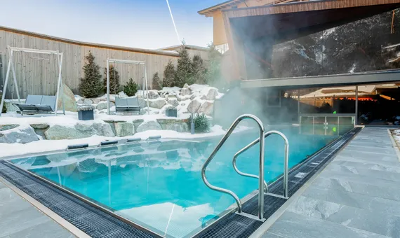 Beheizter Außenpool mit aufsteigendem Dampf, umgeben von Schnee und modernem Alpenhotel