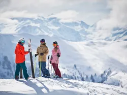 Three skiers in colorful gear standing on a snowy mountain with alpine peaks in the background