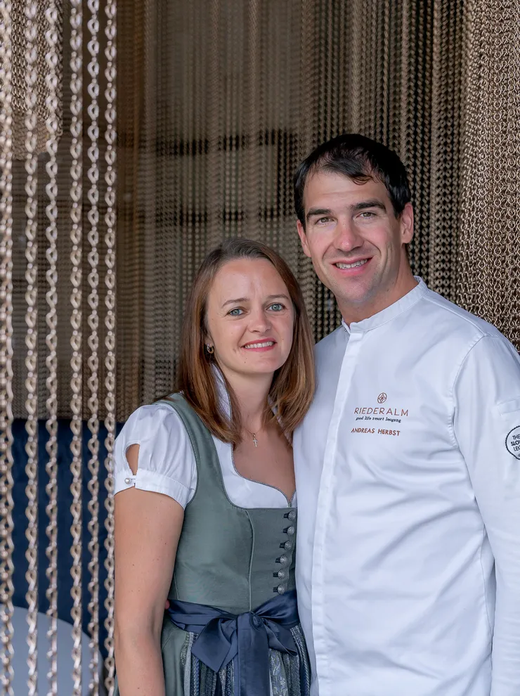 Couple in front of chain curtain, man in Riederalm chef uniform, woman in traditional dirndl dress
