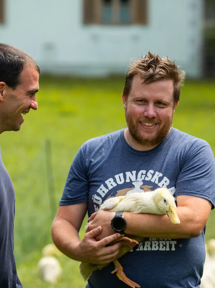 Man holding a duck on a farm while smiling with another man outdoors