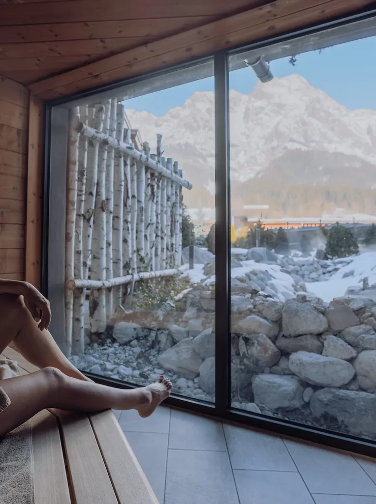 Woman relaxing in sauna with scenic snowy mountain view through large glass window