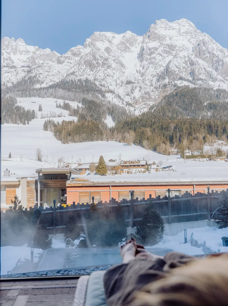 Person relaxing indoors with view of snowy mountains and alpine village through large window