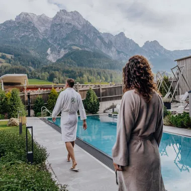 Paar im Bademantel geht am Außenpool eines Wellnesshotels in den Alpen entlang