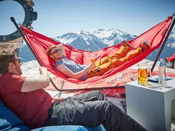 Two men relaxing in hammocks on snow with drinks and scenic mountain view in the background