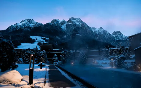 Beheizter Außenpool im verschneiten Bergresort bei Sonnenuntergang mit Alpenpanorama