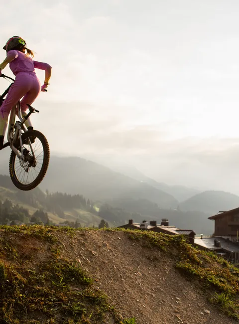 Mountainbikerin in pinkem Outfit springt auf Dirt-Trail bei Sonnenuntergang nahe Alpendorf