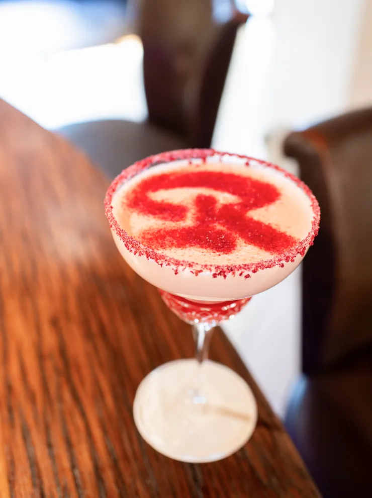 Red and white cocktail in a coupe glass with sugar rim and letter R dusted on top
