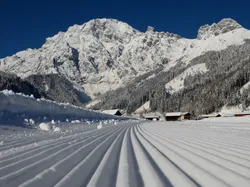 Präparierte Langlaufloipe mit schneebedeckten Alpen und Hütten im Hintergrund