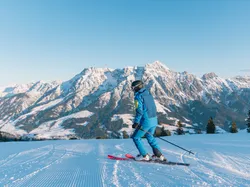Skifahrer in blauer Ausrüstung auf präparierter Piste vor verschneitem Alpenpanorama