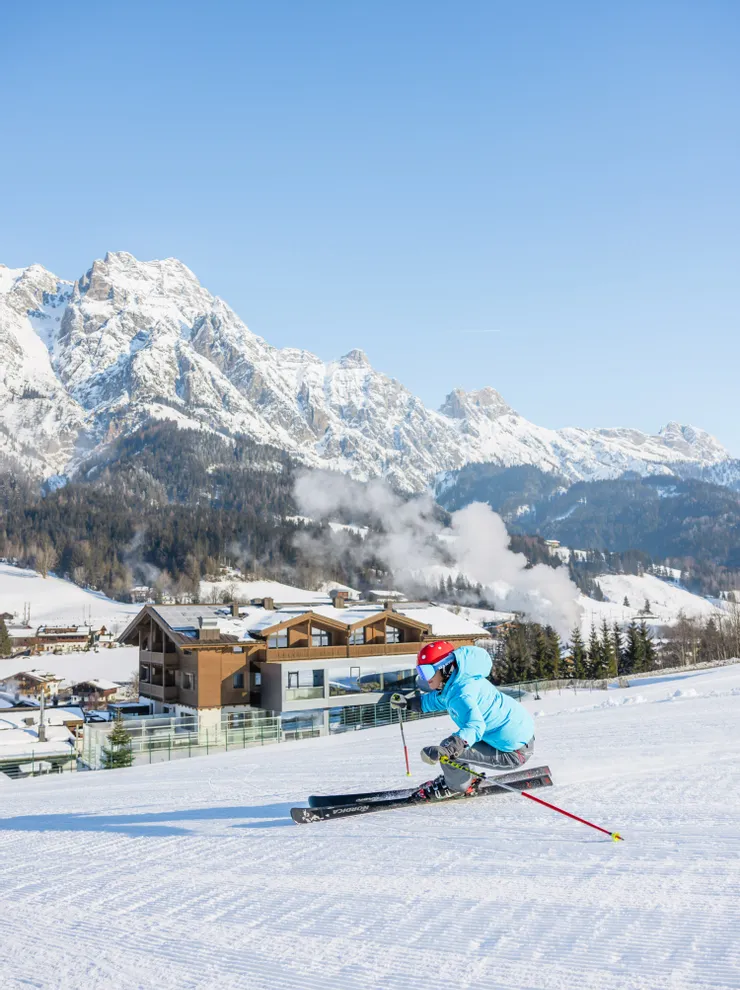 Skifahrer in blauer Jacke carvt eine präparierte Piste mit schneebedeckten Alpen im Hintergrund hinunter