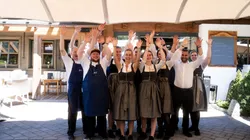 Lächelndes Restaurantteam in traditioneller bayerischer Tracht hebt draußen die Hände