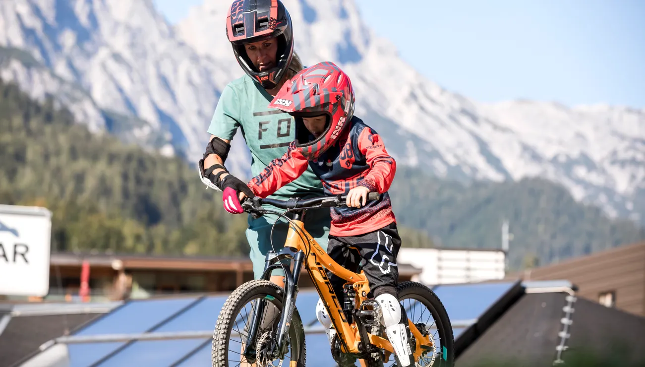 Adult teaching child mountain biking on trail with scenic alpine background