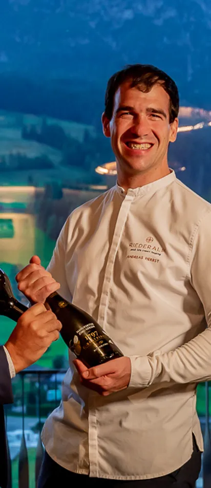 Three men in fine dining restaurant holding wine bottles with mountain view in background