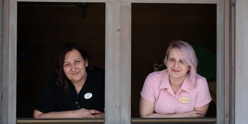 Zwei Mitarbeiterinnen in Uniform lehnen sich lächelnd aus einem Holzfenster