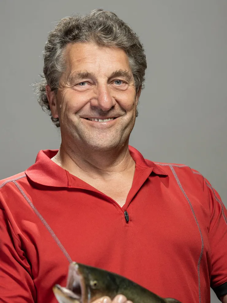 Older man in red shirt holding a fresh fish and smiling in front of gray background