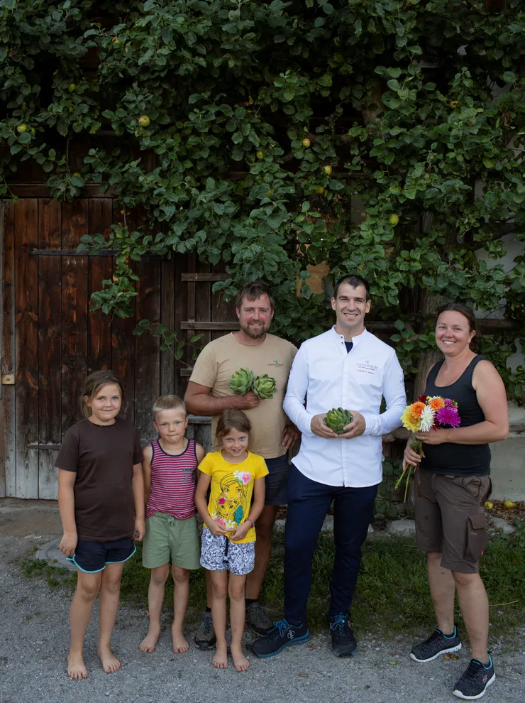 Family with chef holding artichokes and flowers in front of rustic wooden house with green foliage
