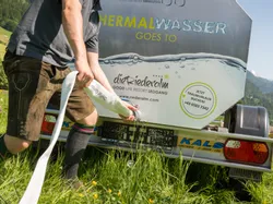 Mann in Lederhose schließt Schlauch an Thermalwasser-Tankanhänger im Grünen an