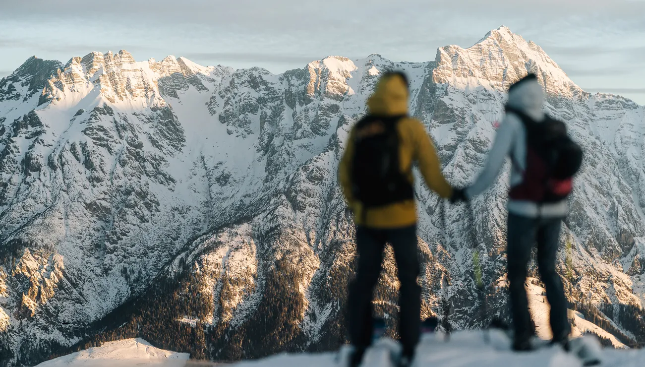 Two hikers in winter gear overlooking snow-covered Alpine mountains at sunrise